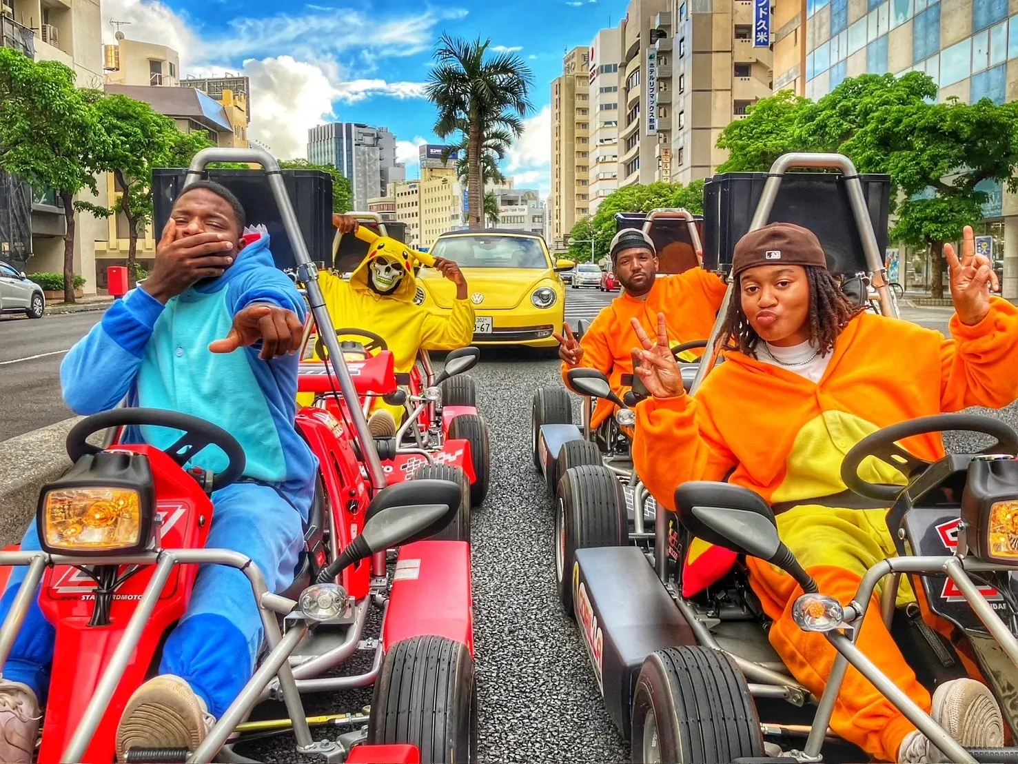 Four riders in cosplay at the wheel on a wide Naha avenue under a palm and a blue sky