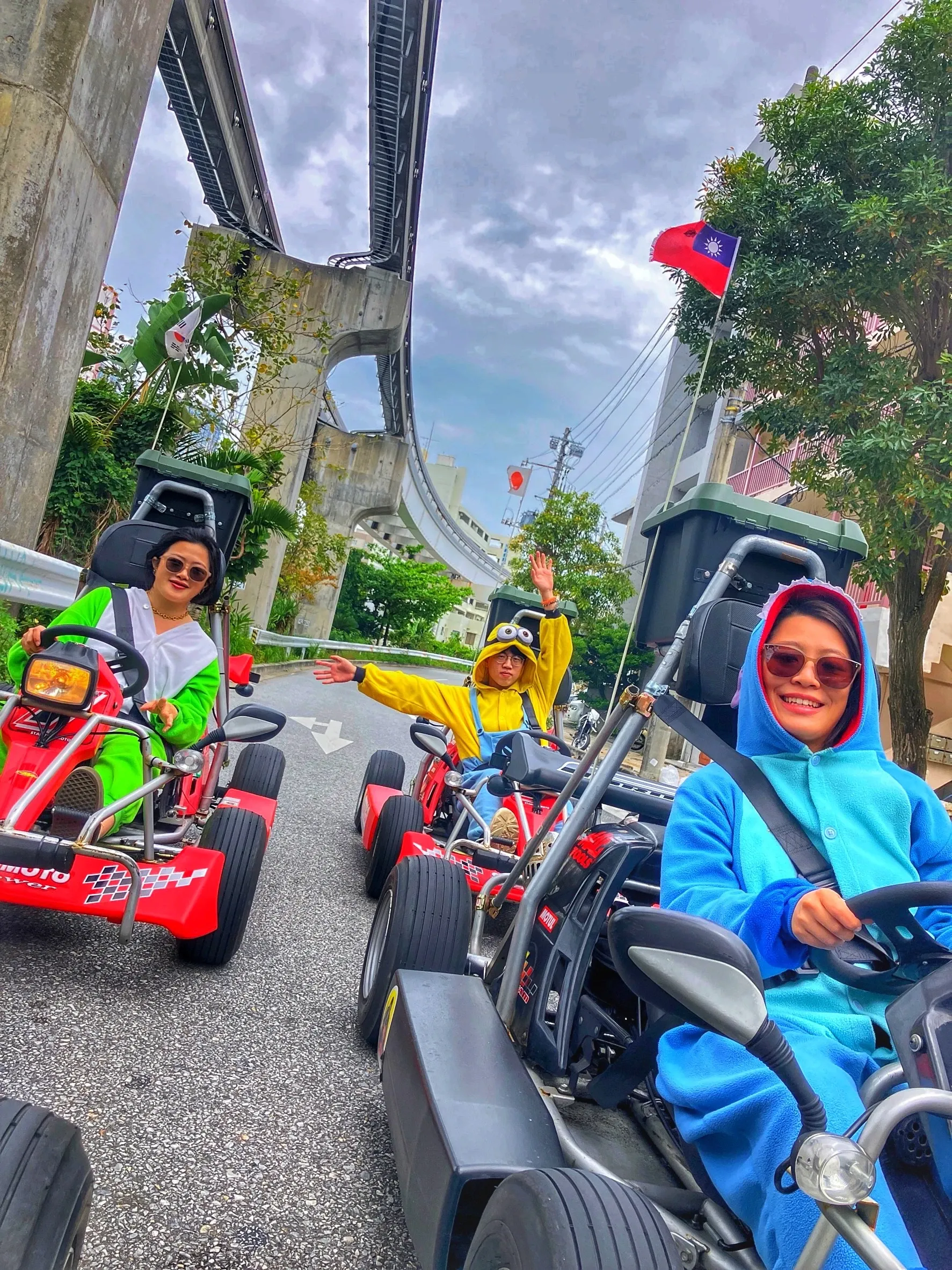 Three go karts on a curving road beneath the monorail guideway, riders in green, yellow and blue kigurumi waving