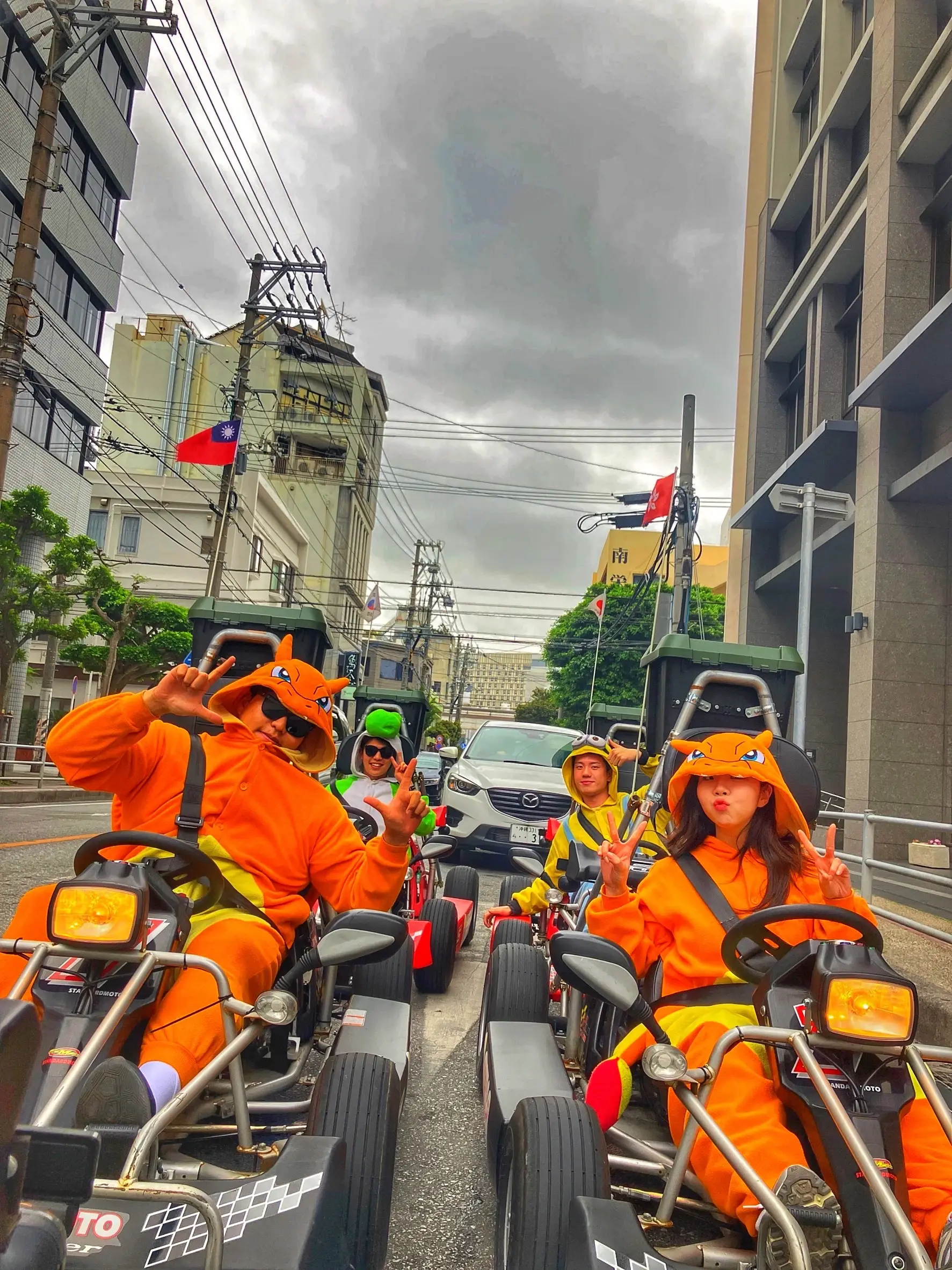 Riders in matching orange kigurumi at the wheel on a narrow Naha street, a car waiting behind the convoy