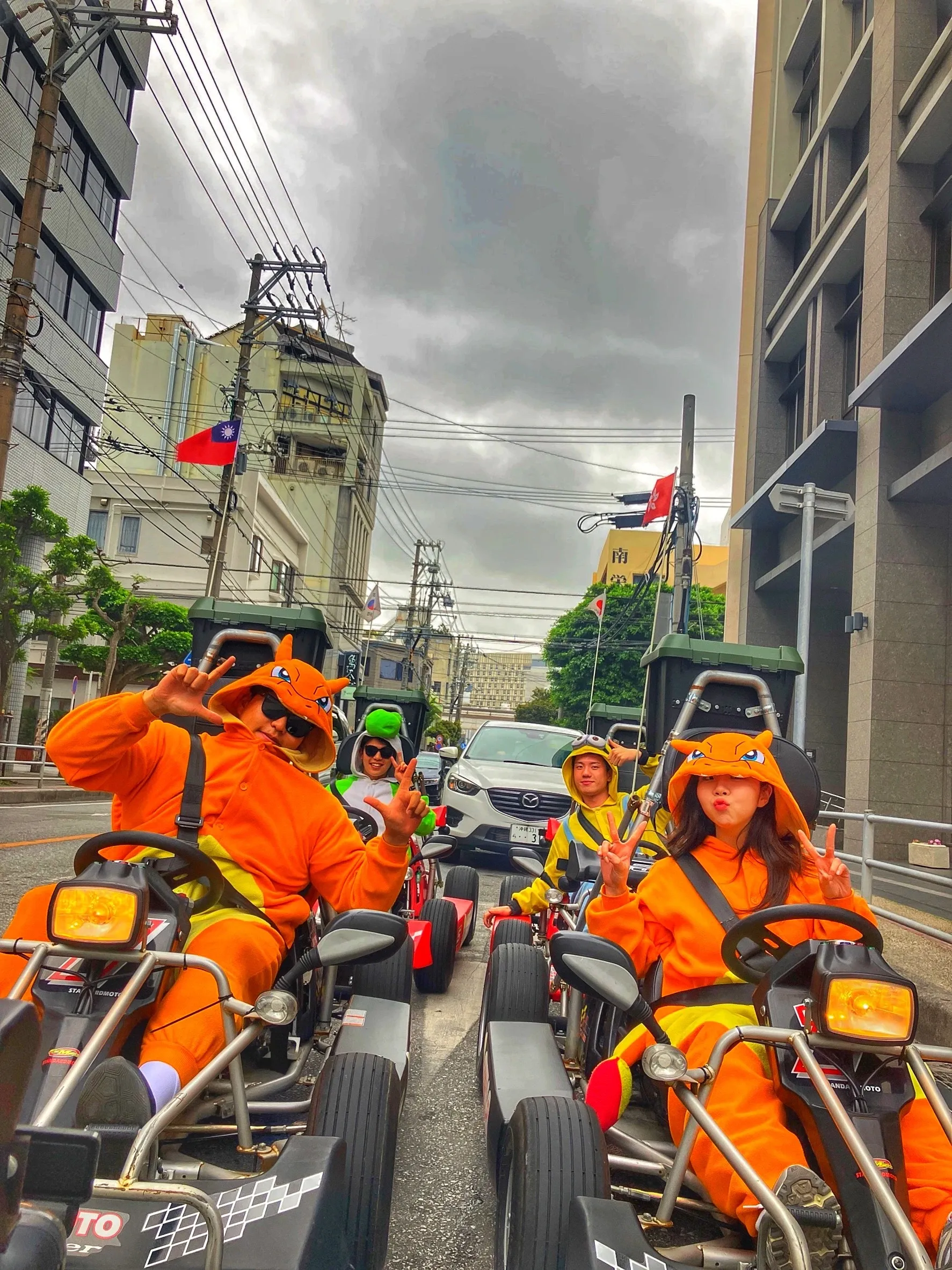 Riders in matching orange kigurumi at the wheel on a narrow Naha street, a car waiting behind the convoy