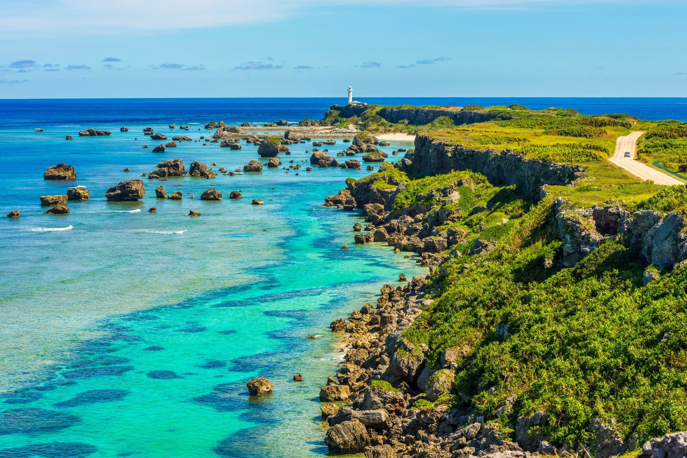 A white lighthouse on a green cape above turquoise coral shallows on the Okinawa coast