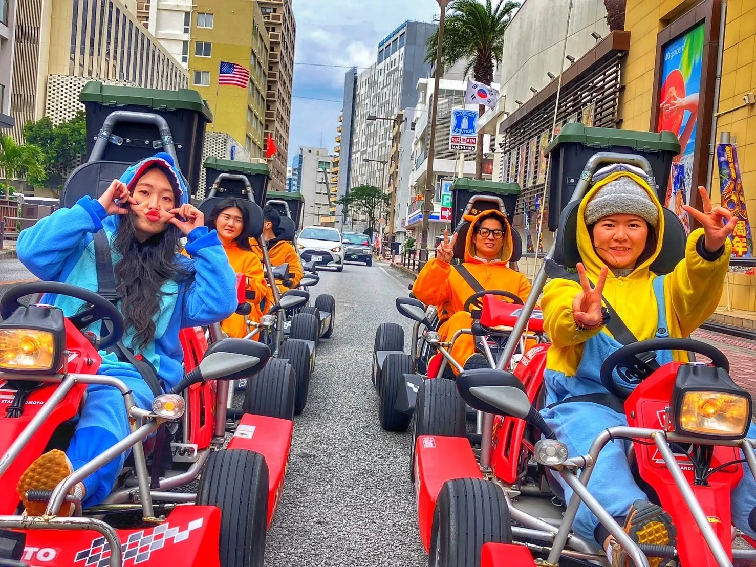 A group of riders in blue, orange and yellow kigurumi at the wheel of karts on Kokusai Dori, flags and shopfronts above them