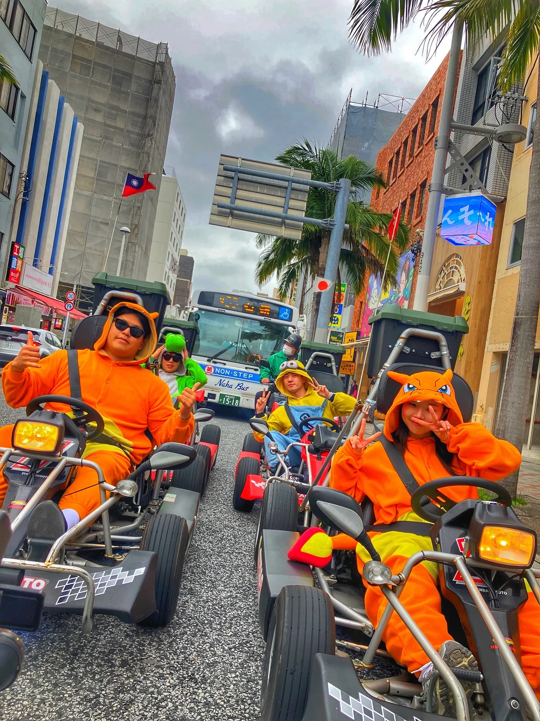 Riders in matching orange kigurumi at the wheel of go karts on a Naha shopping street, a city bus and palms ahead of them