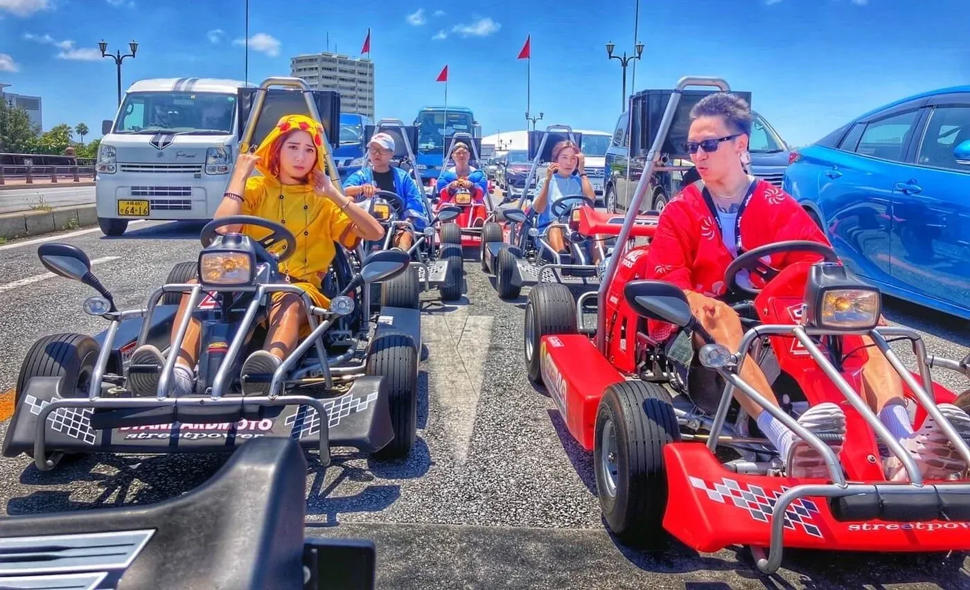 Riders waving from their karts at a junction under a bright blue sky